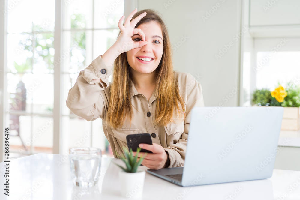 Beautiful young woman using smartphone and computer with happy face smiling doing ok sign with hand on eye looking through fingers Beautiful young woman using smartphone and computer with happy face smiling doing ok sign with hand on eye looking through fingers