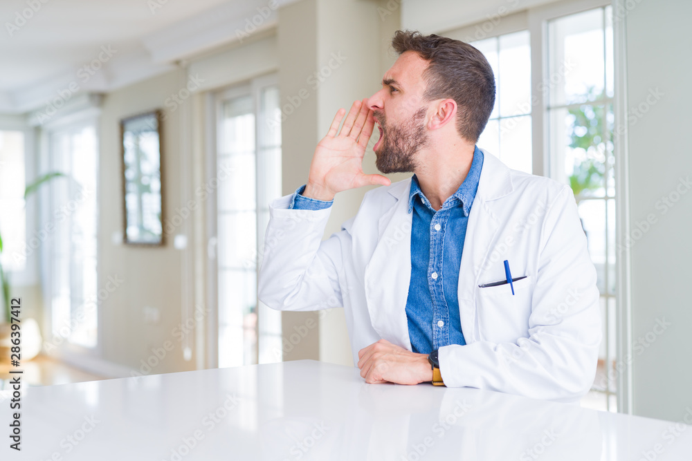 Handsome doctor man wearing medical coat at the clinic shouting and screaming loud to side with hand on mouth. Communication concept. Handsome doctor man wearing medical coat at the clinic shouting and screaming loud to side with hand on mouth. Communication concept.