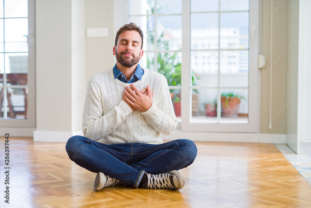 Handsome man wearing casual sweater sitting on the floor at home smiling with hands on chest with closed eyes and grateful gesture on face. Health concept.
