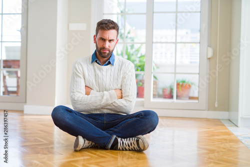 Handsome man wearing casual sweater sitting on the floor at home skeptic and nervous, disapproving expression on face with crossed arms. Negative person.