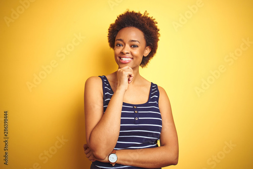 Beauitul african american woman wearing summer t-shirt over isolated yellow background looking confident at the camera smiling with crossed arms and hand raised on chin. Thinking positive.