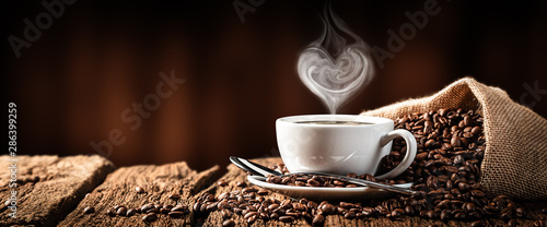 Biała filiżanka gorącej kawy z parą w kształcie serca na starym wyblakłym stole z jutowym workiem i fasolą