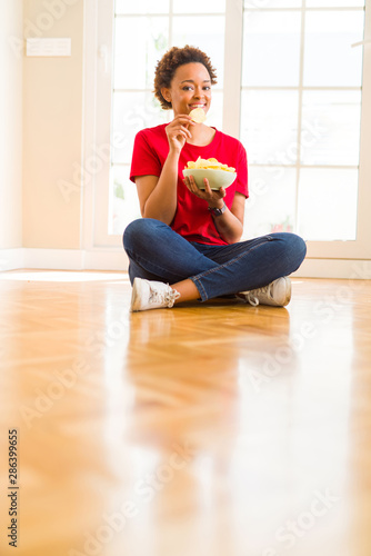 Young beautiful african american woman with afro hair eating chips sitting on the floor at home