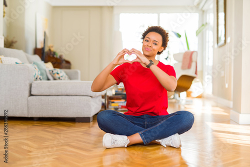 Young beautiful african american woman sitting on the floor at home smiling in love showing heart symbol and shape with hands. Romantic concept.