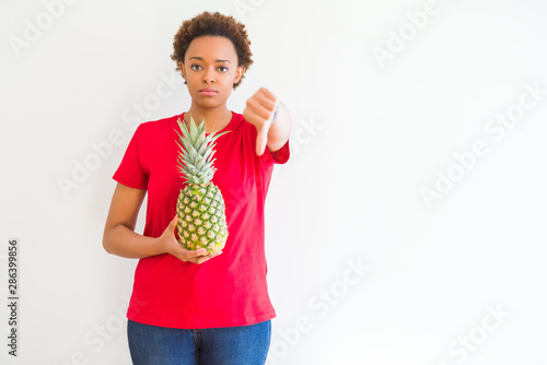 Young african american woman holding fresh healthy pineapple fruit with angry face, negative sign showing dislike with thumbs down, rejection concept