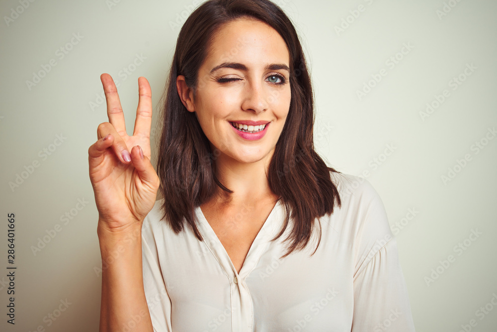 Young beautiful woman wearing casual shirt standing over white isolated background smiling with happy face winking at the camera doing victory sign. Number two. Young beautiful woman wearing casual shirt standing over white isolated background smiling with happy face winking at the camera doing victory sign. Number two.