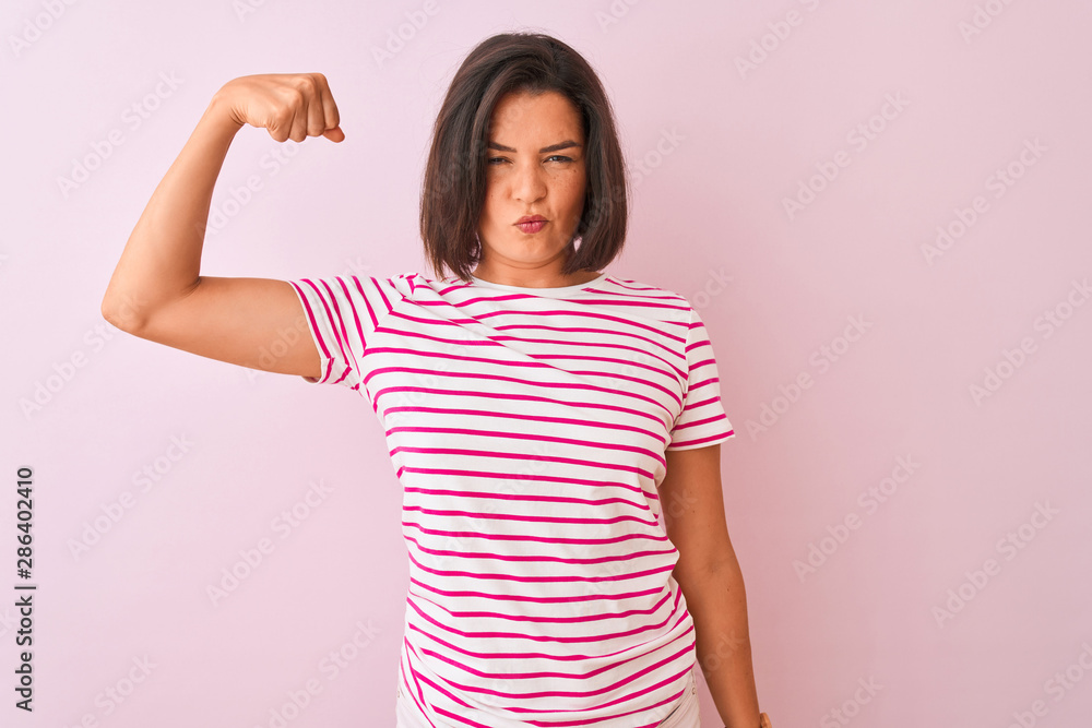 Young beautiful woman wearing striped t-shirt standing over isolated pink background Strong person showing arm muscle, confident and proud of power