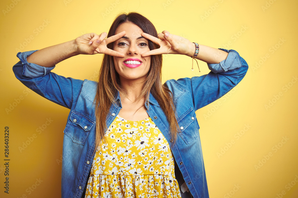 Young beautiful woman standing over yellow isolated background Doing peace symbol with fingers over face, smiling cheerful showing victory Young beautiful woman standing over yellow isolated background Doing peace symbol with fingers over face, smiling cheerful showing victory