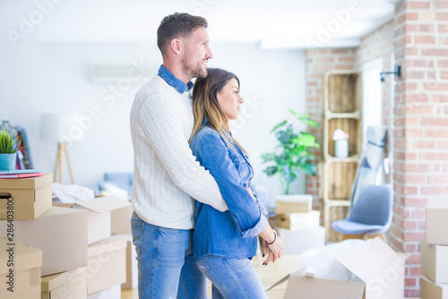 Young beautiful couple standing at new home around cardboard boxes looking to side, relax profile pose with natural face with confident smile.