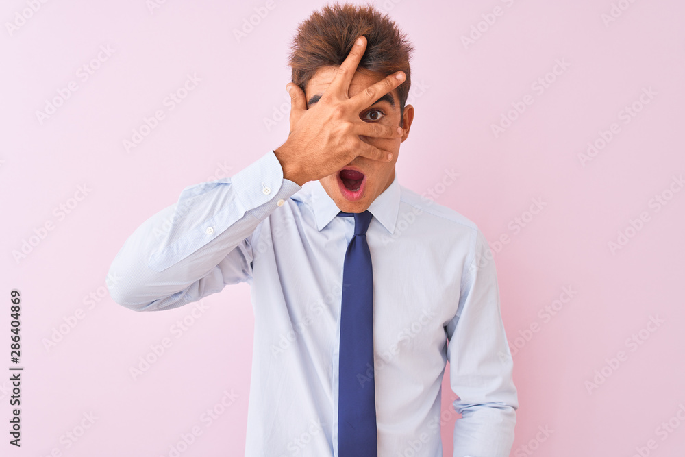 Young handsome businessman wearing shirt and tie standing over isolated pink background peeking in shock covering face and eyes with hand, looking through fingers with embarrassed expression.