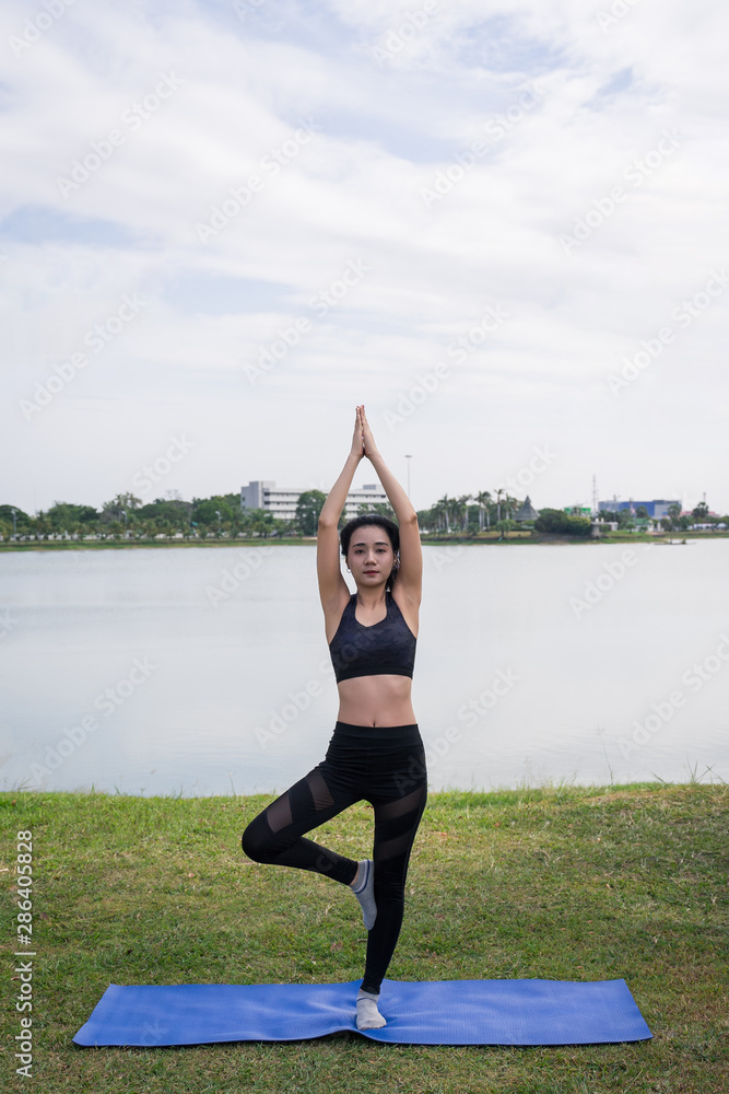 Fototapeta premium Slim beautiful women practices yoga poses on the mat beside a lake at the outdoor park with green grass background. Yoga and life health concept with copy and paste space.