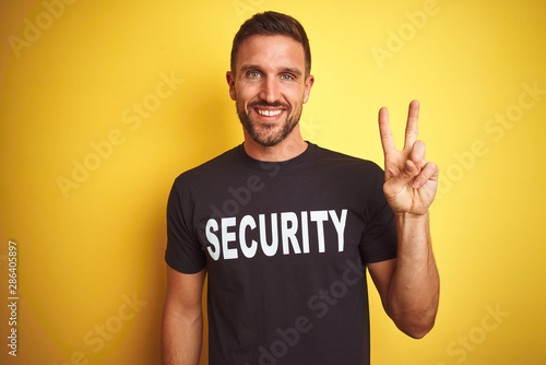 Young safeguard man wearing security uniform over yellow isolated background smiling with happy face winking at the camera doing victory sign. Number two. © Krakenimages.com