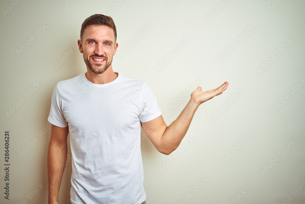Young handsome man wearing casual white t-shirt over isolated background smiling cheerful presenting and pointing with palm of hand looking at the camera. Young handsome man wearing casual white t-shirt over isolated background smiling cheerful presenting and pointing with palm of hand looking at the camera.
