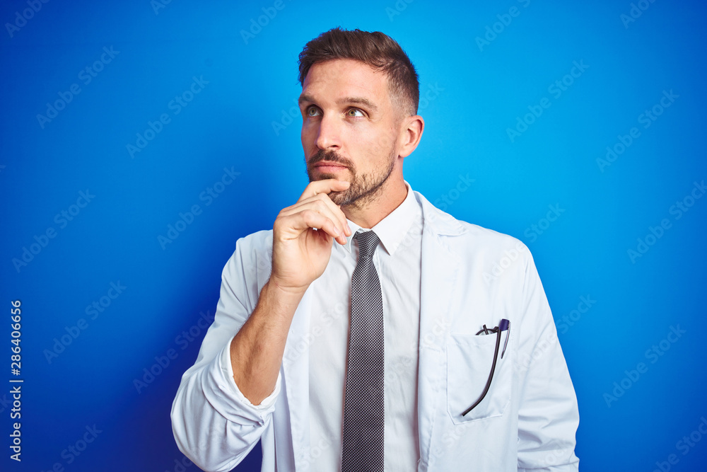 Young handsome doctor man wearing white professional coat over isolated background with hand on chin thinking about question, pensive expression. Smiling with thoughtful face. Doubt concept. Young handsome doctor man wearing white professional coat over isolated background with hand on chin thinking about question, pensive expression. Smiling with thoughtful face. Doubt concept.