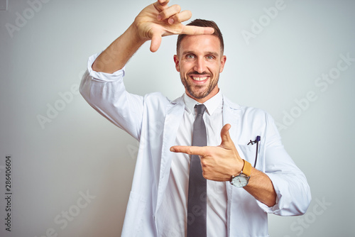 Young handsome doctor man wearing white profressional coat over isolated background smiling making frame with hands and fingers with happy face. Creativity and photography concept. photo