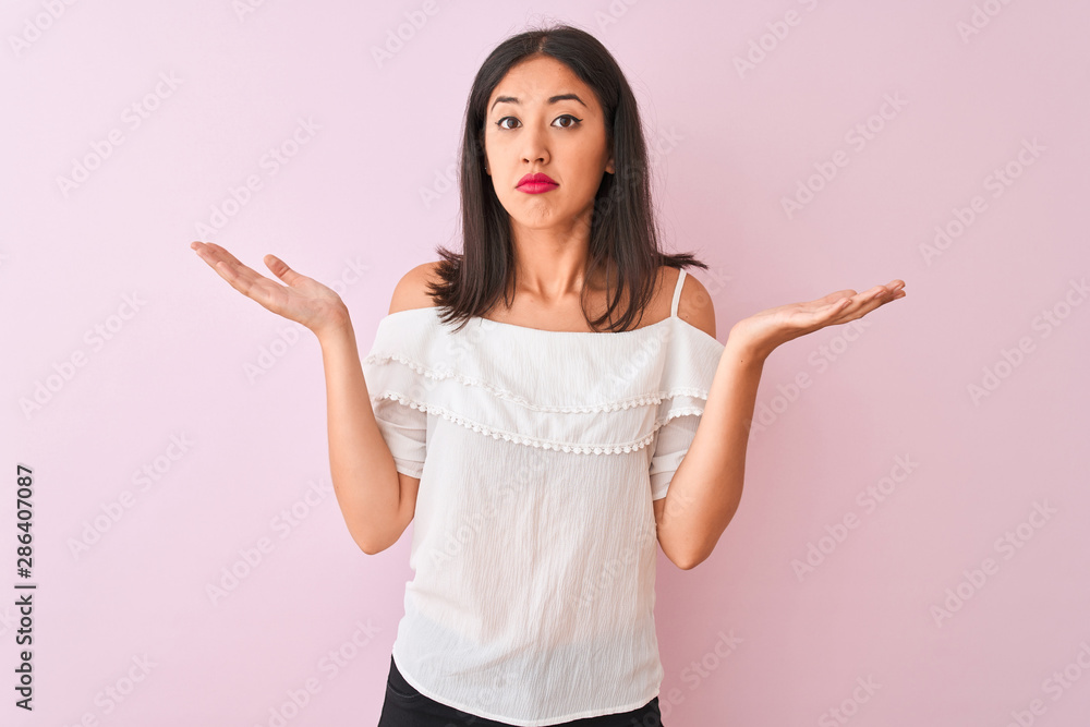 Beautiful chinese woman wearing white t-shirt standing over isolated pink background clueless and confused expression with arms and hands raised. Doubt concept. Beautiful chinese woman wearing white t-shirt standing over isolated pink background clueless and confused expression with arms and hands raised. Doubt concept.
