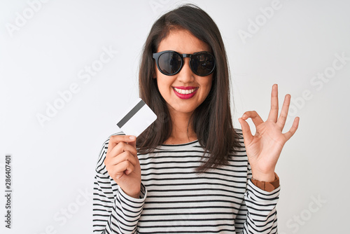 Young chinese woman wearing sunglasses holding credit card over isolated white background doing ok sign with fingers, excellent symbol
