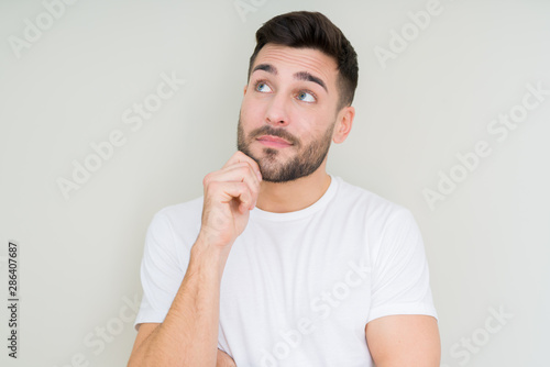 Young handsome man wearing casual white t-shirt over isolated background with hand on chin thinking about question, pensive expression. Smiling with thoughtful face. Doubt concept.