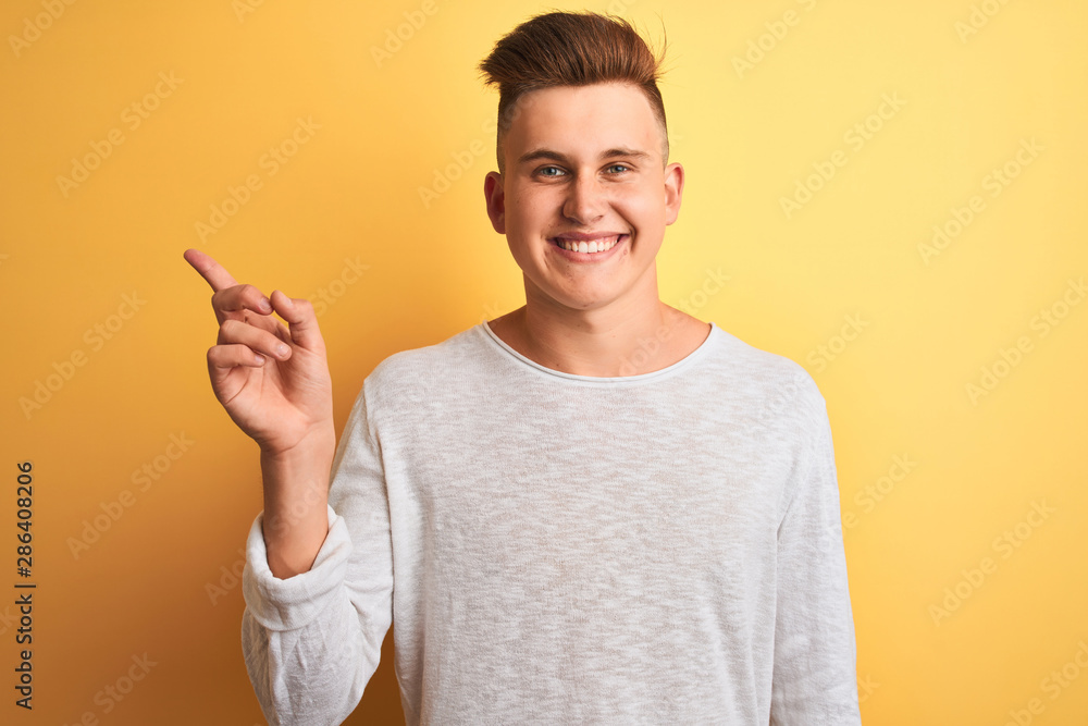 Young handsome man wearing white casual t-shirt standing over isolated yellow background with a big smile on face, pointing with hand and finger to the side looking at the camera.