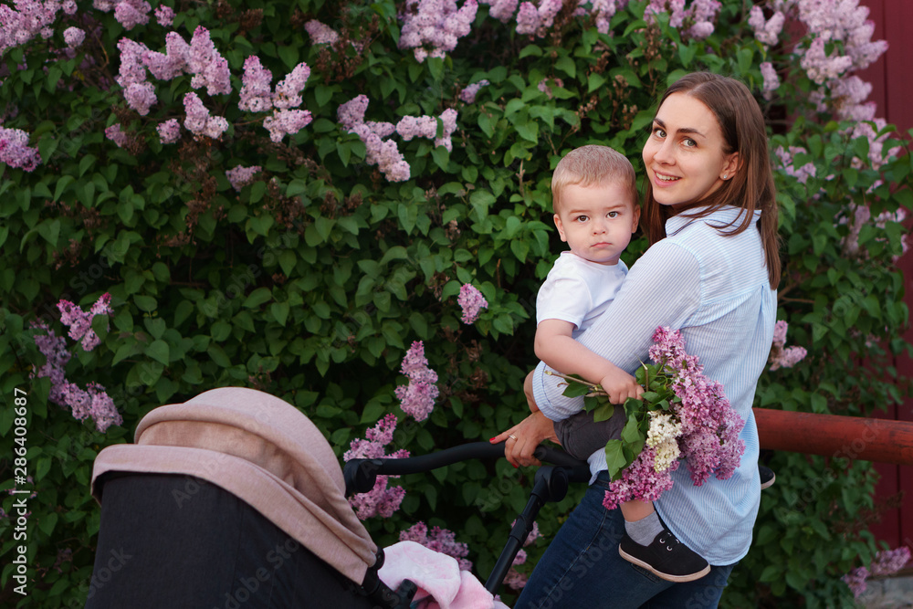 Young caring mother on walk with two children on background of spring flowering. Eldest child in her arms is sitting in her mothers arms. Baby is sleeping in stroller. Happiness to be parent. 