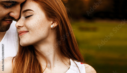 Close up portrait of a amazing couple while woman with red hair and freckles is leaning head with closed eyes smiling on her boyfriend outside.