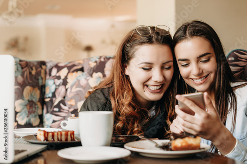 Lovely plus size female with red hair having fun time laughing with her female friend while looking at smartphone in a cafe.