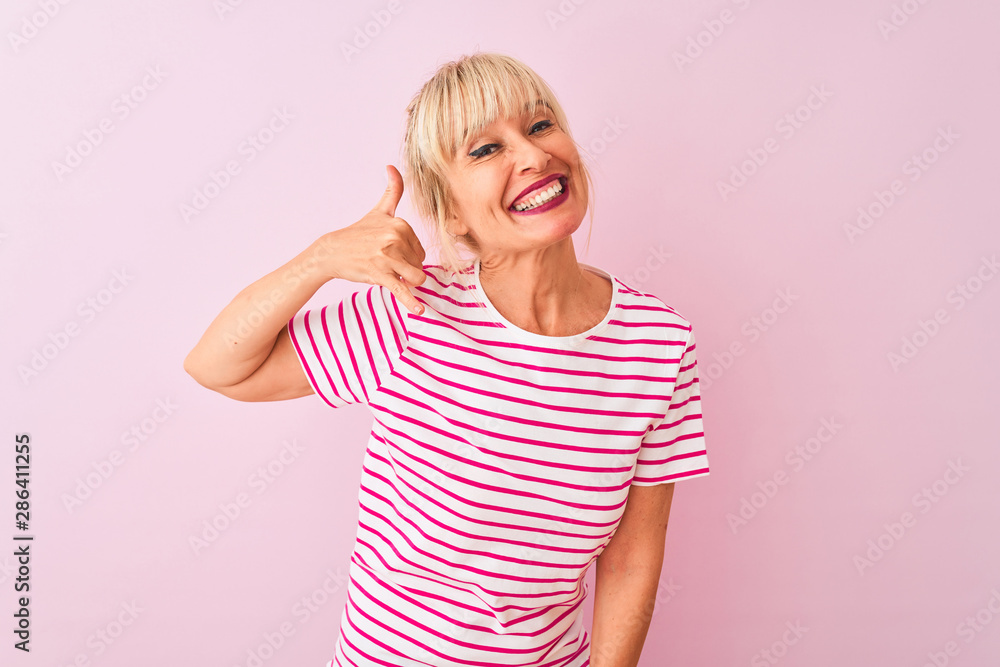 Middle age woman wearing striped t-shirt standing over isolated pink background smiling doing phone gesture with hand and fingers like talking on the telephone. Communicating concepts. Middle age woman wearing striped t-shirt standing over isolated pink background smiling doing phone gesture with hand and fingers like talking on the telephone. Communicating concepts.