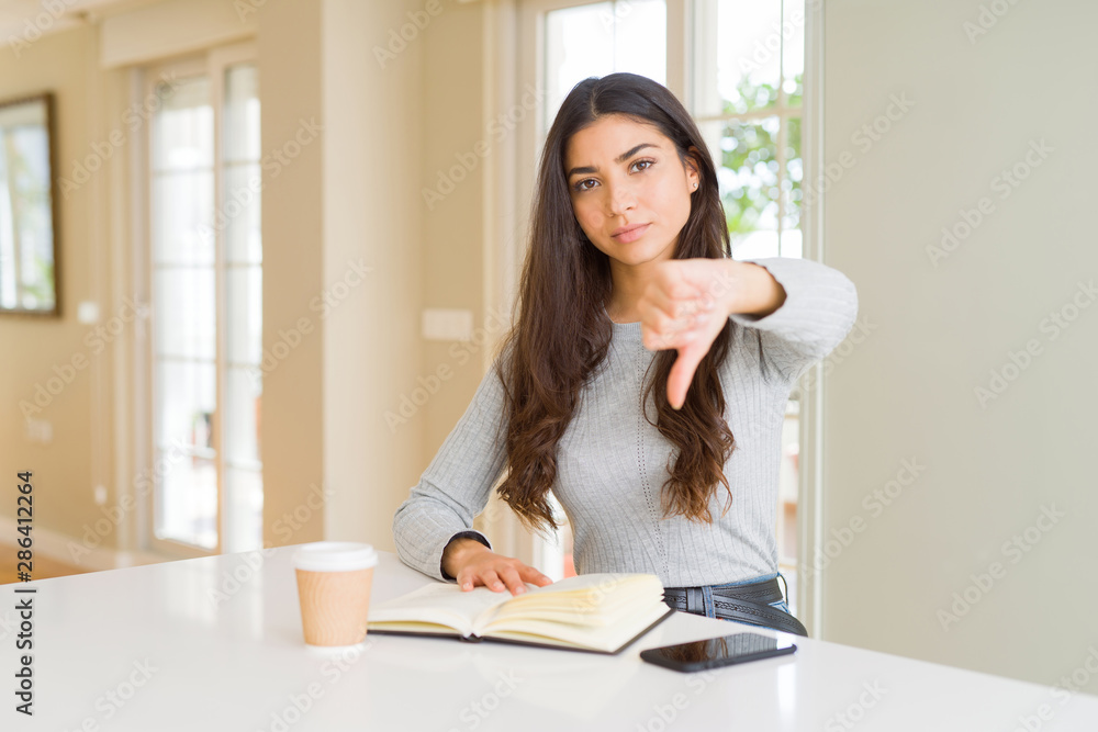 Young woman reading a book and drinking a coffe with angry face, negative sign showing dislike with thumbs down, rejection concept Young woman reading a book and drinking a coffe with angry face, negative sign showing dislike with thumbs down, rejection concept