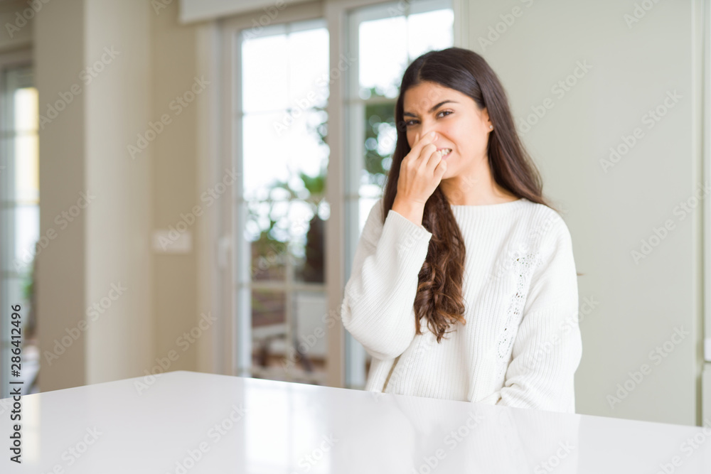 Young beautiful woman at home on white table smelling something stinky and disgusting, intolerable smell, holding breath with fingers on nose. Bad smells concept. Young beautiful woman at home on white table smelling something stinky and disgusting, intolerable smell, holding breath with fingers on nose. Bad smells concept.
