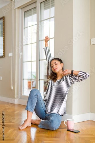 Beautiful young woman stretching and yawning in the morning sitting on the floor drinking a cup of coffee for breakfast