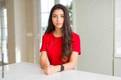 Young beautiful woman at home on white table with serious expression on face. Simple and natural looking at the camera.