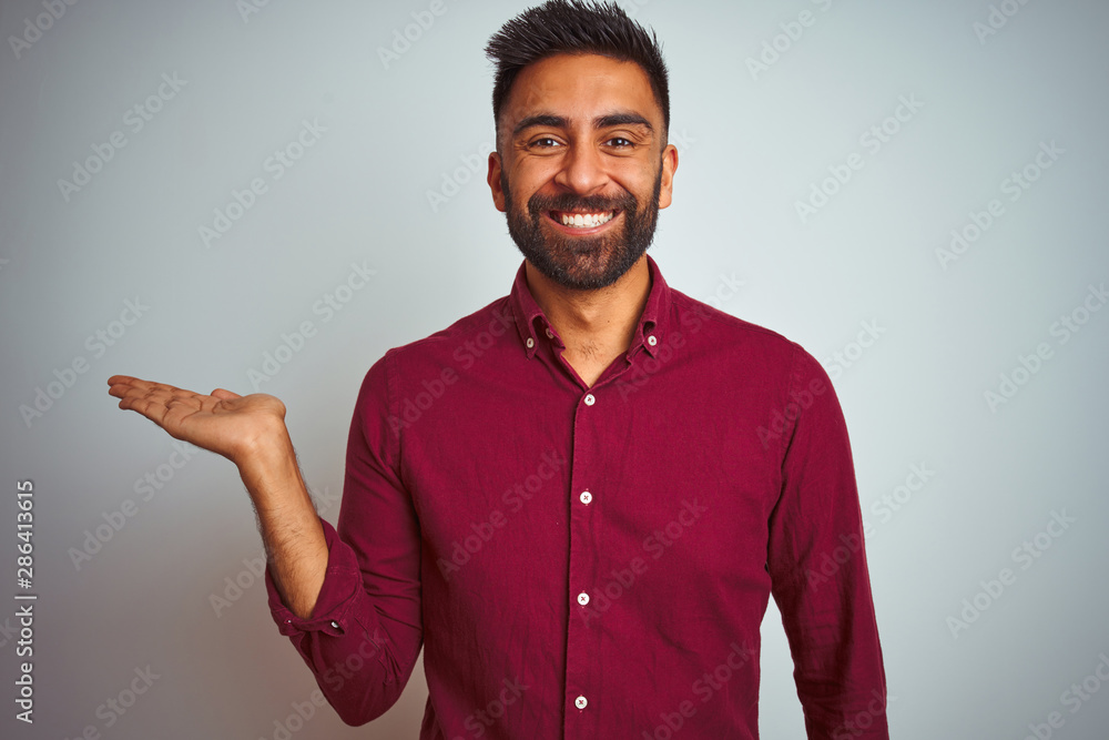 Young indian man wearing red elegant shirt standing over isolated grey background smiling cheerful presenting and pointing with palm of hand looking at the camera. Young indian man wearing red elegant shirt standing over isolated grey background smiling cheerful presenting and pointing with palm of hand looking at the camera.