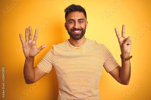 Young indian man wearing t-shirt standing over isolated yellow background showing and pointing up with fingers number seven while smiling confident and happy.