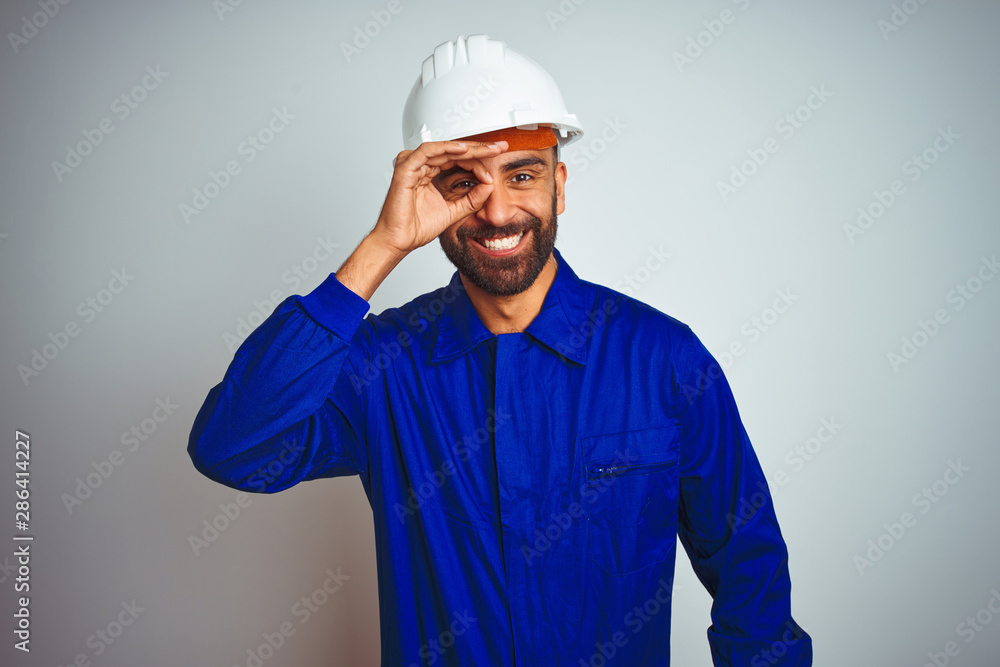 Handsome indian worker man wearing uniform and helmet over isolated white background doing ok gesture with hand smiling, eye looking through fingers with happy face.