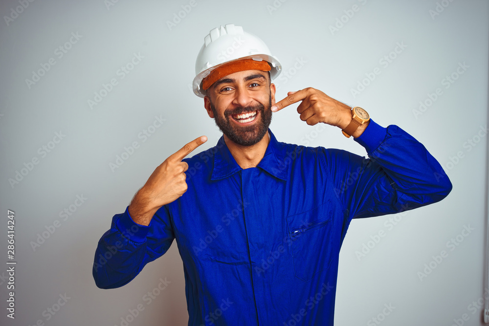 Handsome indian worker man wearing uniform and helmet over isolated white background smiling cheerful showing and pointing with fingers teeth and mouth. Dental health concept.