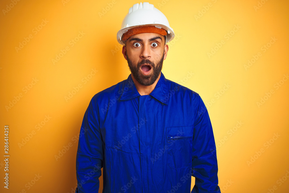 Handsome indian worker man wearing uniform and helmet over isolated yellow background afraid and shocked with surprise expression, fear and excited face.