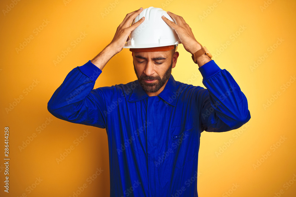 Handsome indian worker man wearing uniform and helmet over isolated yellow background suffering from headache desperate and stressed because pain and migraine. Hands on head.