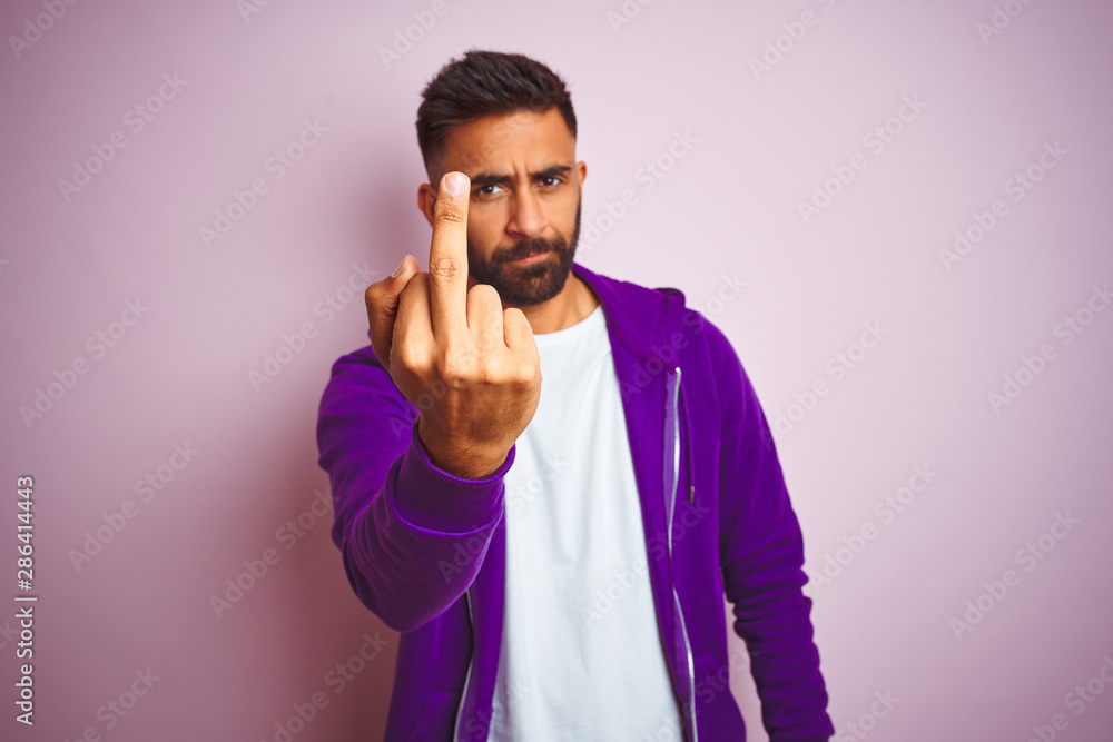 Young indian man wearing purple sweatshirt standing over isolated pink background Showing middle finger, impolite and rude fuck off expression Young indian man wearing purple sweatshirt standing over isolated pink background Showing middle finger, impolite and rude fuck off expression