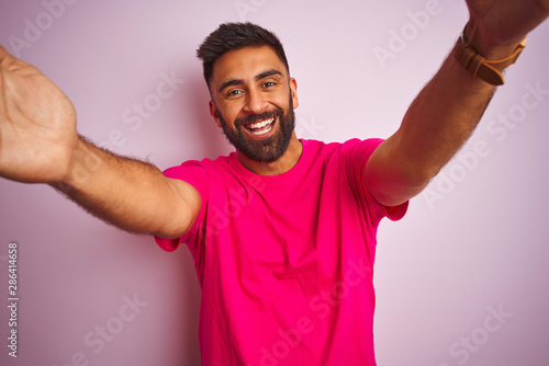 Young indian man wearing t-shirt standing over isolated pink background looking at the camera smiling with open arms for hug. Cheerful expression embracing happiness.