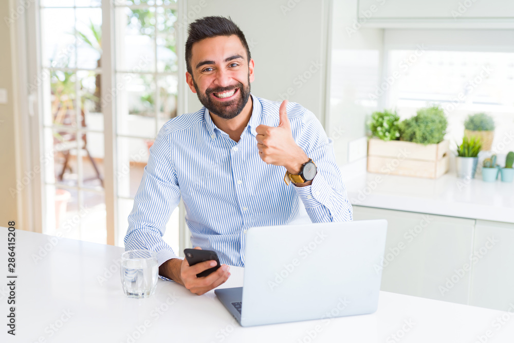 Handsome hispanic business man using laptop and smartphone happy with big smile doing ok sign, thumb up with fingers, excellent sign