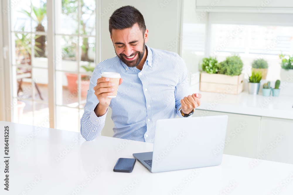 Handsome hispanic man working using computer laptop and drinking a cup of coffee screaming proud and celebrating victory and success very excited, cheering emotion