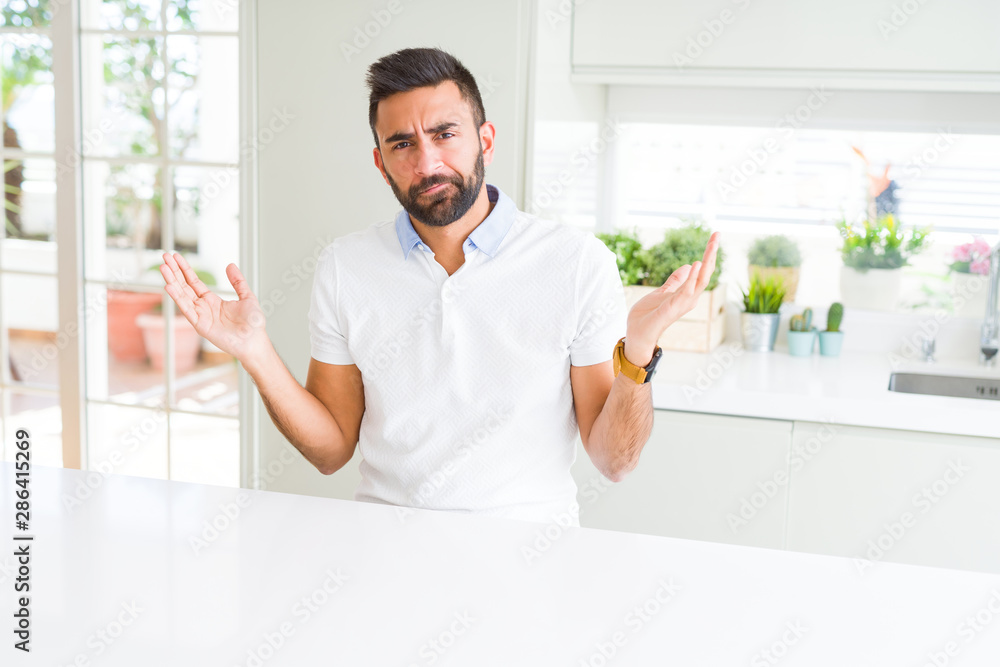 Handsome hispanic man casual white t-shirt at home clueless and confused expression with arms and hands raised. Doubt concept.