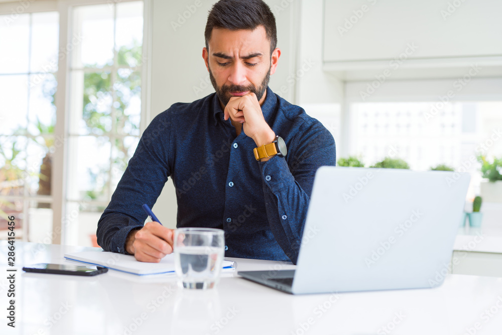 Fototapeta premium Handsome hispanic man working using computer and writing on a paper serious face thinking about question, very confused idea
