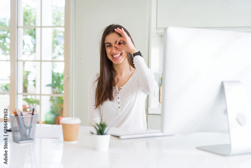 Beautiful young woman working using computer doing ok gesture with hand smiling, eye looking through fingers with happy face. Beautiful young woman working using computer doing ok gesture with hand smiling, eye looking through fingers with happy face.