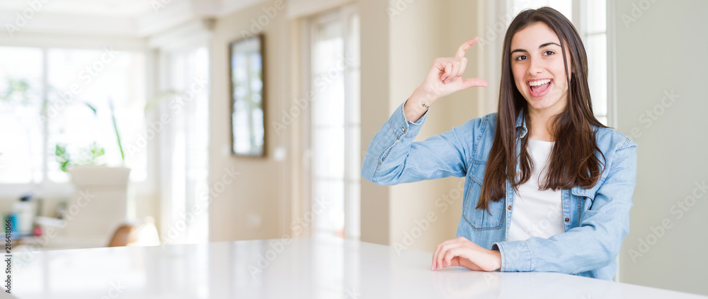 Wide angle picture of beautiful young woman sitting on white table at home smiling and confident gesturing with hand doing size sign with fingers while looking and the camera. Measure concept. Wide angle picture of beautiful young woman sitting on white table at home smiling and confident gesturing with hand doing size sign with fingers while looking and the camera. Measure concept.