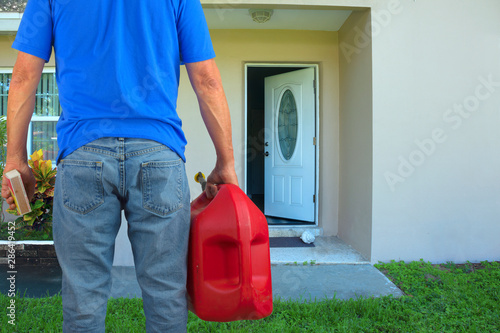 Arsonist man with red plastic gasoline can container and box of striking matches preparing to commit arson crime and maliciously and  intentionally burn down a house with an open front door.