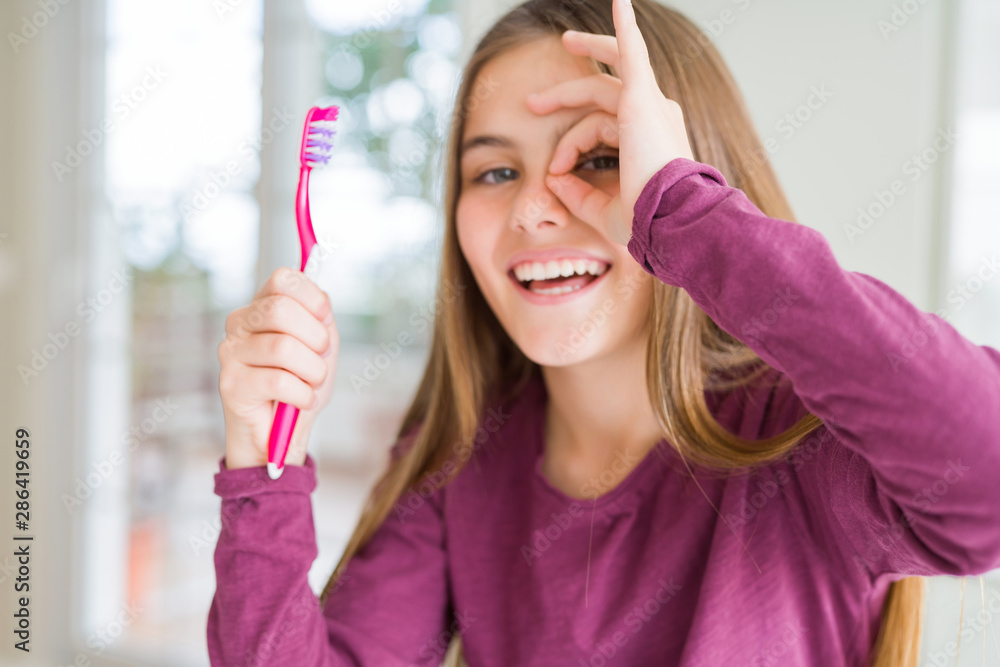Beautiful young girl kid holding pink dental toothbrush with happy face smiling doing ok sign with hand on eye looking through fingers Beautiful young girl kid holding pink dental toothbrush with happy face smiling doing ok sign with hand on eye looking through fingers