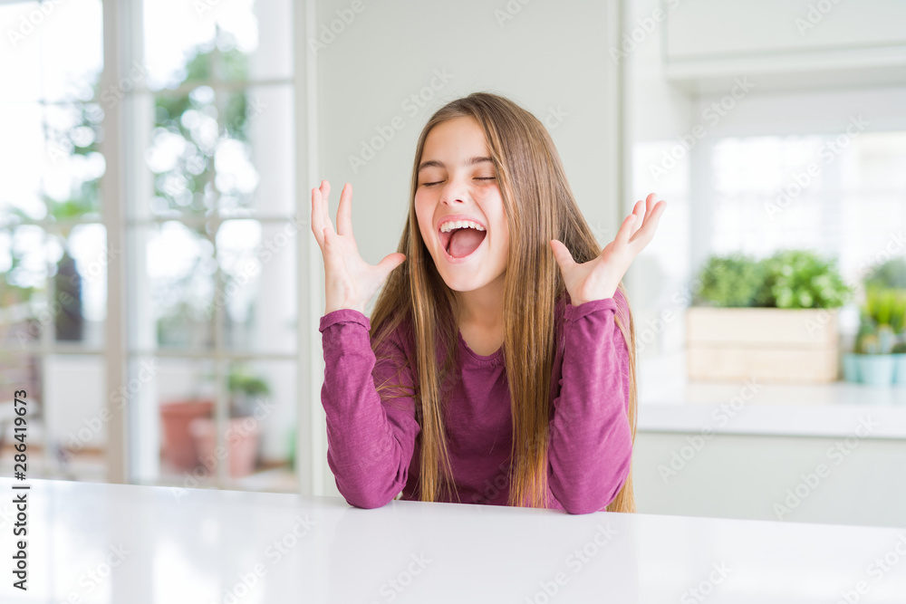 Beautiful young girl kid on white table celebrating mad and crazy for success with arms raised and closed eyes screaming excited. Winner concept Beautiful young girl kid on white table celebrating mad and crazy for success with arms raised and closed eyes screaming excited. Winner concept
