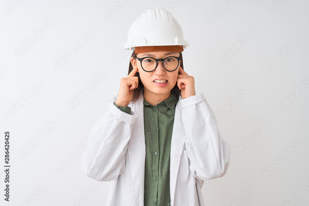 Young chinese engineer woman wearing coat helmet glasses over isolated white background covering ears with fingers with annoyed expression for the noise of loud music. Deaf concept. Young chinese engineer woman wearing coat helmet glasses over isolated white background covering ears with fingers with annoyed expression for the noise of loud music. Deaf concept.