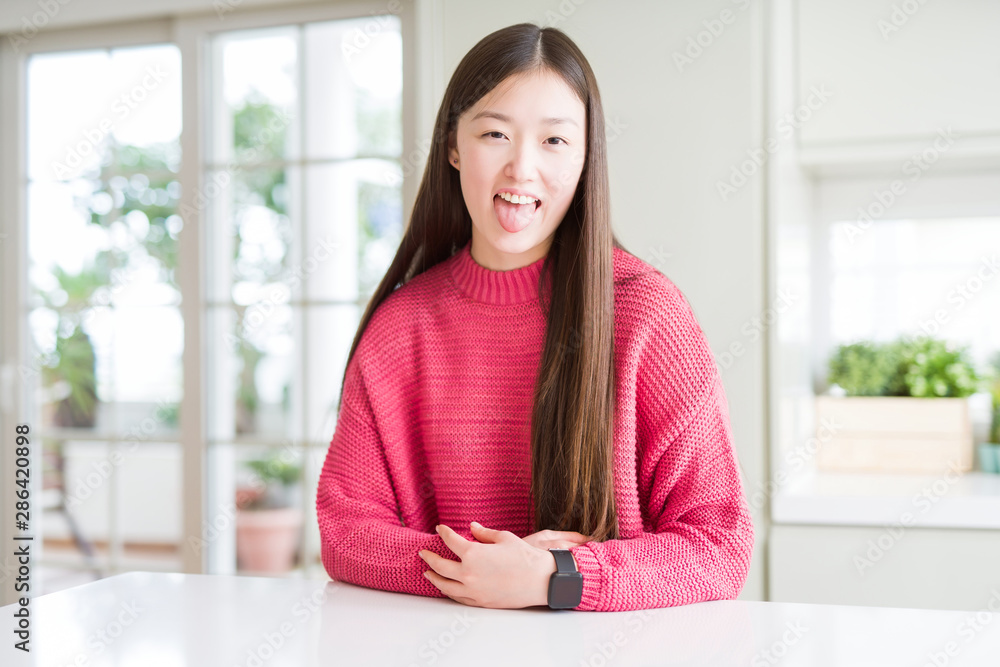 Beautiful Asian woman wearing pink sweater on white table sticking tongue out happy with funny expression. Emotion concept. Beautiful Asian woman wearing pink sweater on white table sticking tongue out happy with funny expression. Emotion concept.